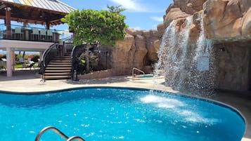 A swimming pool with a waterfall feature, surrounded by rock formations and a covered seating area with lounge chairs.