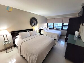 A hotel room with two beds, a large mirror, a desk with a lamp, and a view of the city through the window.