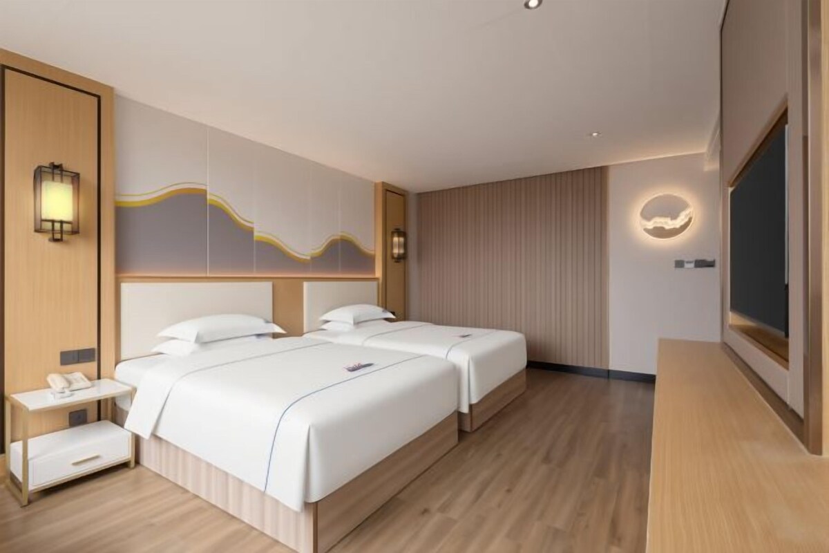Habitación de hotel con dos camas, una mesita de noche, un televisor y una lámpara fijada a la pared.