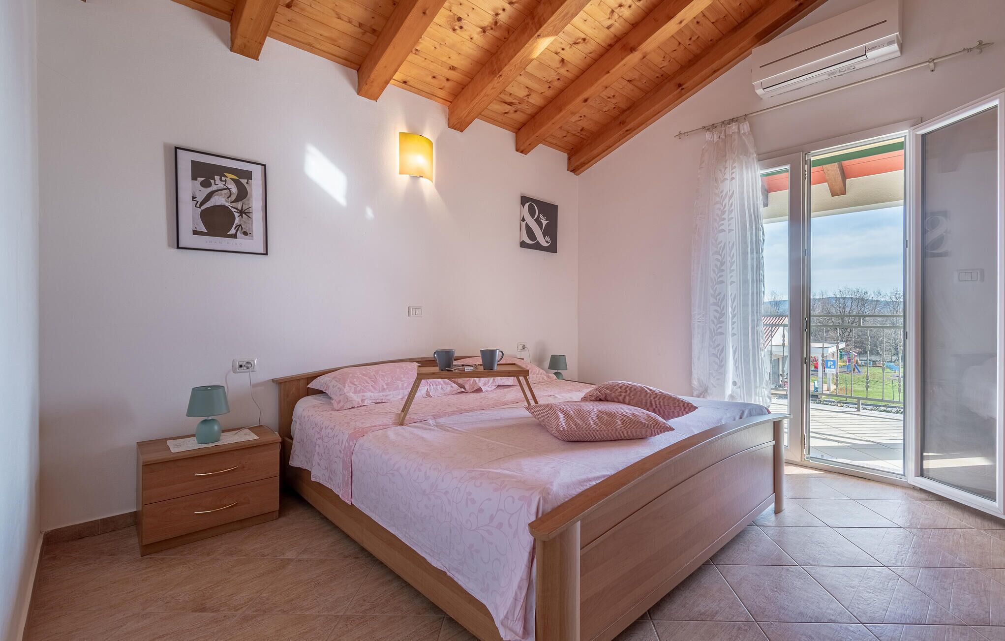 Ein Schlafzimmer mit einem hölzernen Bett, einem Nachttisch, einer Lampe und einem großen Fenster mit Blick nach draußen.