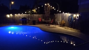 A swimming pool with a blue light at night, surrounded by a wooden fence and outdoor furniture.