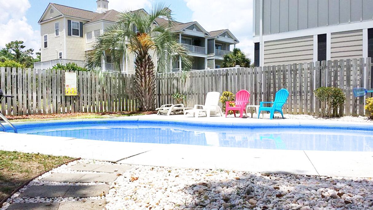 A swimming pool with a wooden fence, a palm tree, and colorful Adirondack chairs.