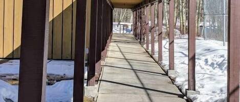 A covered walkway with wooden beams and a concrete path, leading to a building with large windows.