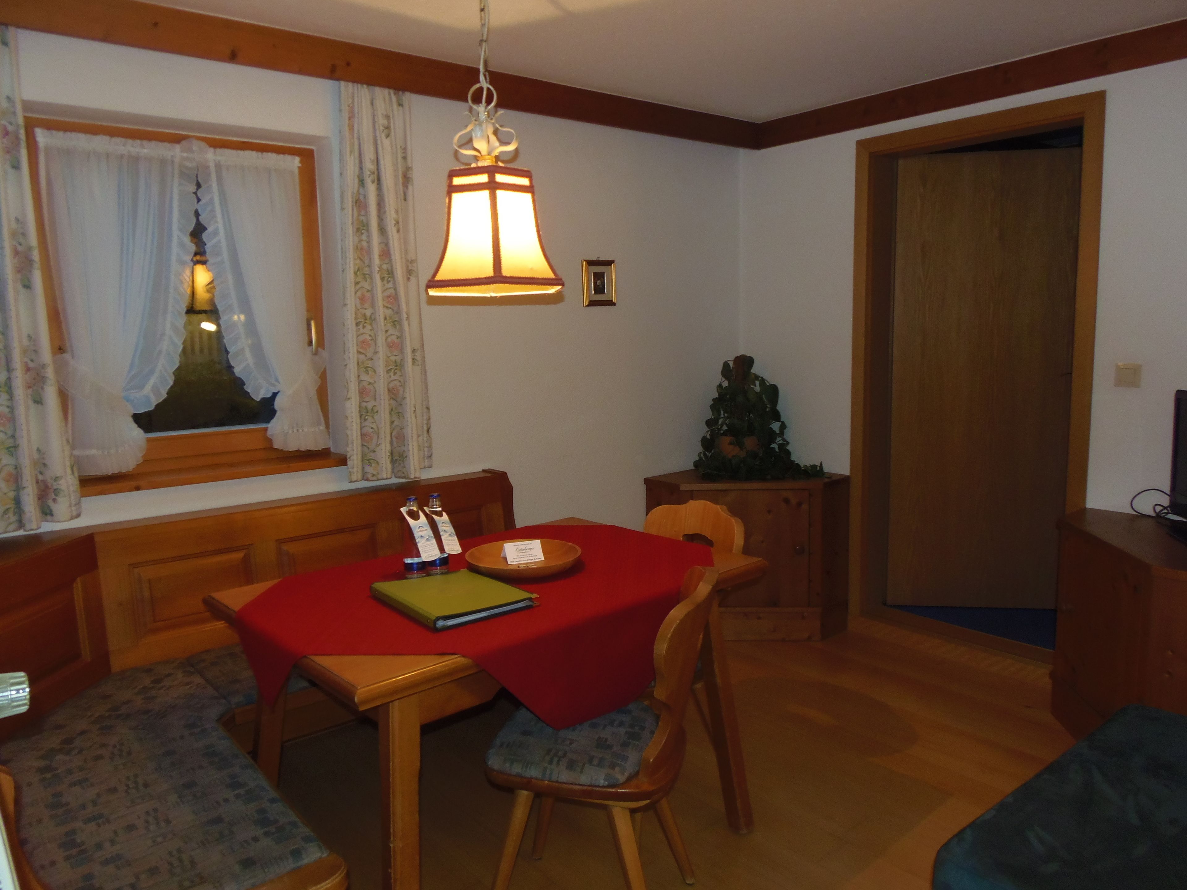 Un coin repas avec une nappe rouge, des chaises en bois, un luminaire suspendu, une fenêtre avec des rideaux et un petit sapin de Noël.