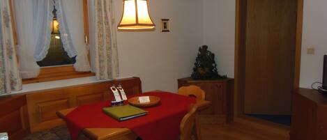 A dining area with a red tablecloth, wooden chairs, a hanging light fixture, a window with curtains, and a small Christmas tree.