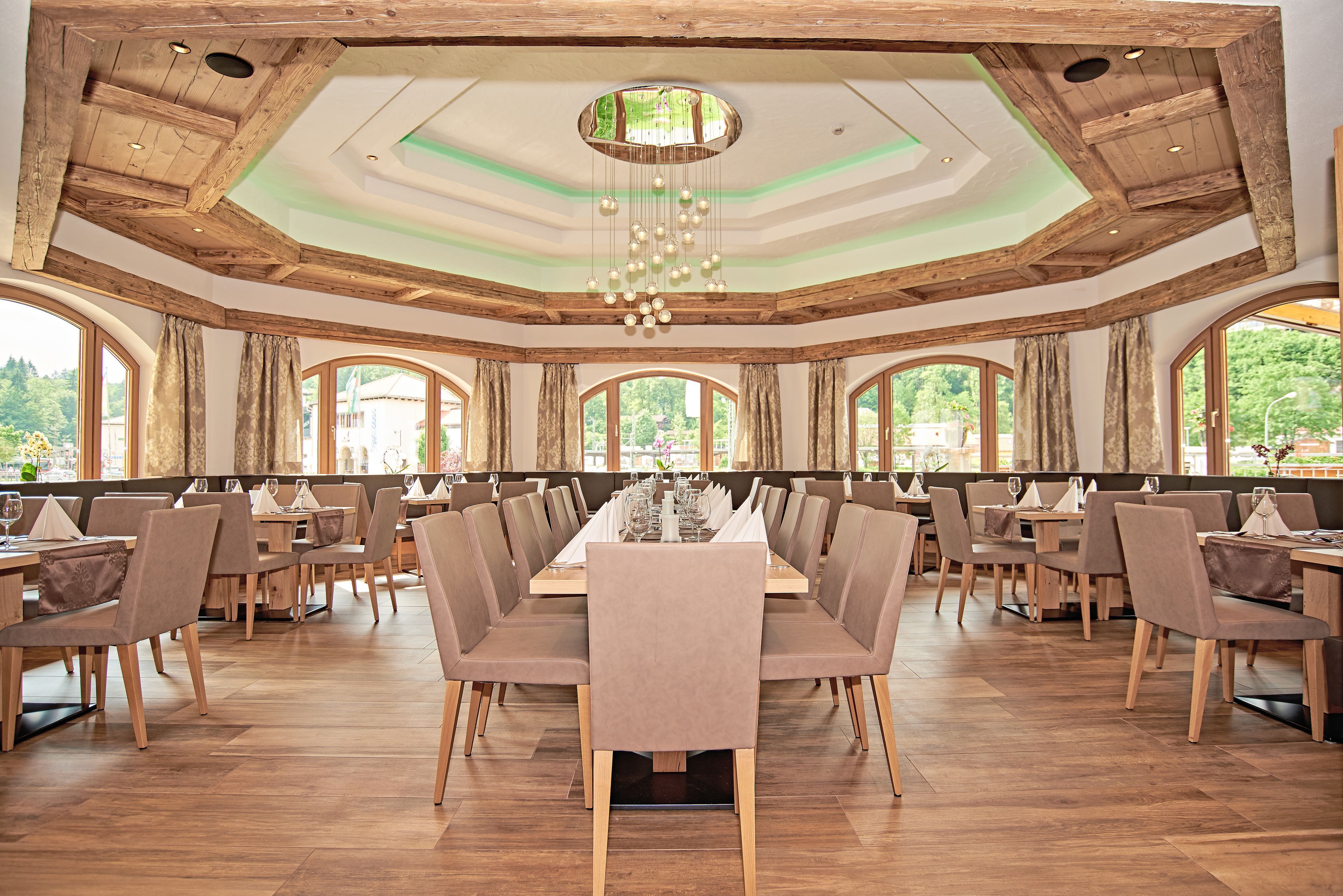 Un amplio comedor con largas mesas dispuestas para muchos invitados, vigas de madera en el techo y grandes ventanales que ofrecen vistas al exterior.