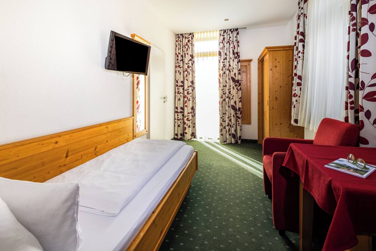 Quarto de hotel com uma cama, televisão fixada na parede, escrivaninha e uma cadeira.