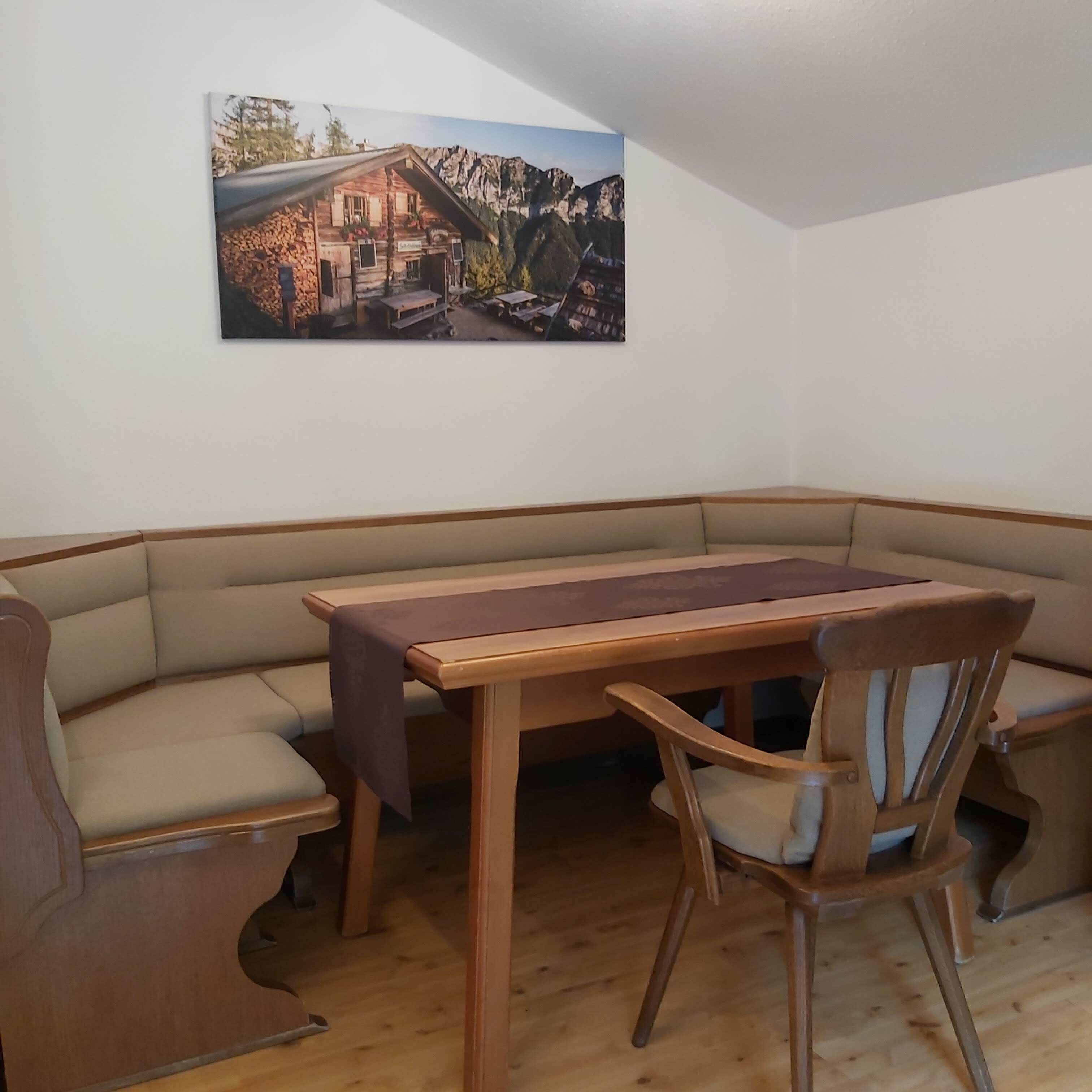 Un coin repas avec une table en bois et deux chaises rembourrées. Un tableau représentant un chalet de montagne est accroché au mur.