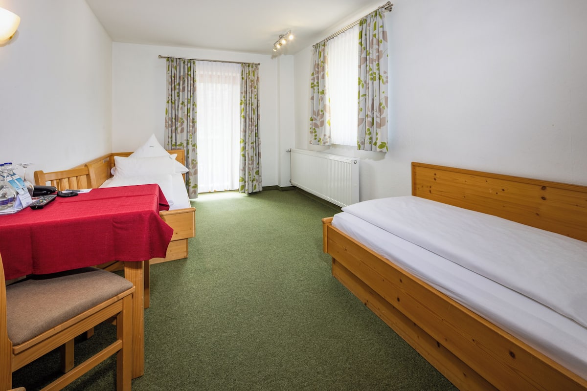 A room with a single bed, a wooden desk with a red tablecloth, a chair, and a window with floral curtains.