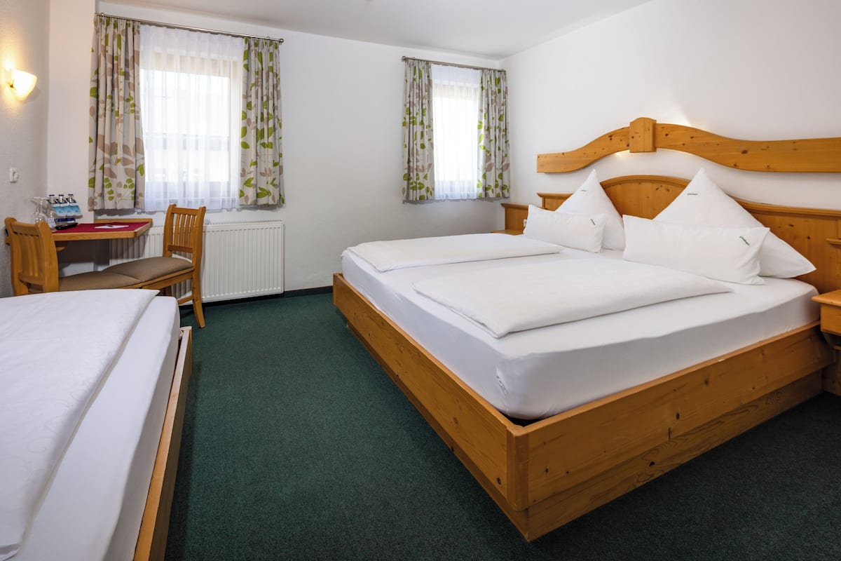 Uma cama de casal com cabeceira de madeira, uma mesinha de cabeceira de madeira, uma escrivaninha com cadeira e duas janelas com cortinas florais.