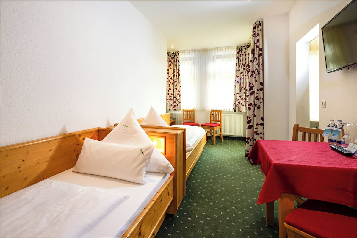 Quarto de hotel com duas camas de solteiro, uma mesa de jantar com cadeiras, uma televisão na parede e uma janela com cortinas.