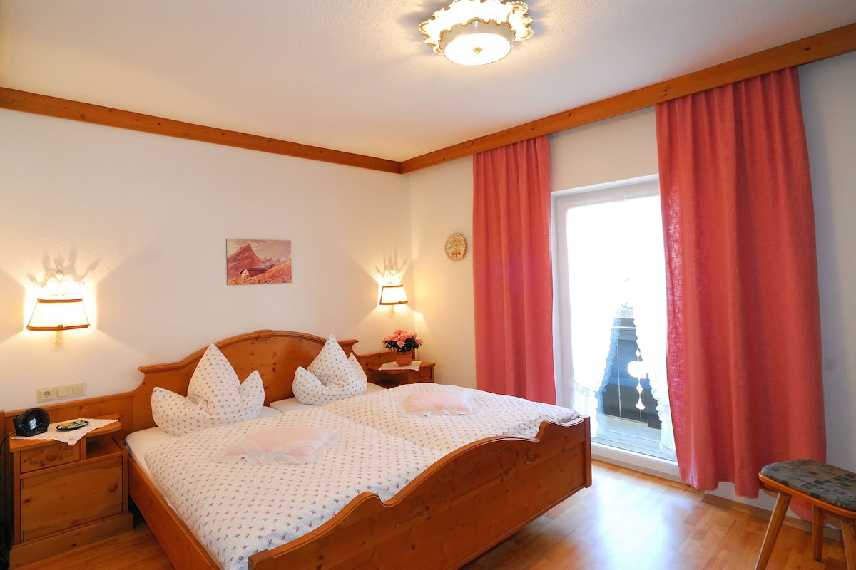 Quarto com cama de madeira, duas luminárias de cabeceira, mesa de cabeceira, janela com cortinas e vista para um edifício.