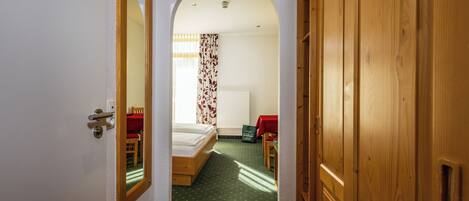 A narrow hallway with a wooden door on the right, a mirror on the left, and a bed with a red bedspread in the background.