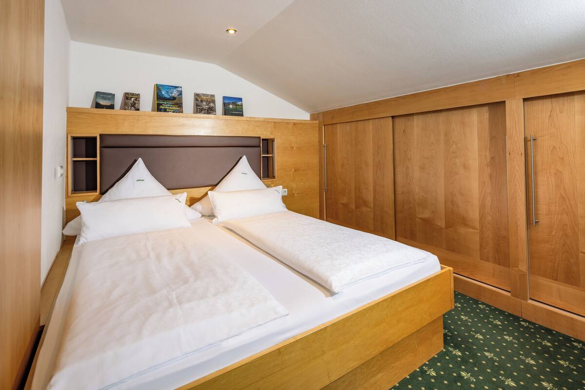 Uma cama de casal com cabeceira de madeira, guarda-roupa de madeira e um tapete verde estampado.