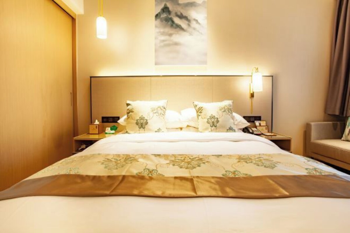 A hotel room with a large bed, a headboard with a light fixture, a bedside table with a lamp, and a painting on the wall.