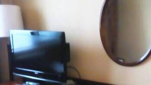 Una habitación con un televisor de pantalla plana sobre un soporte de madera, un espejo redondo en la pared y una mesita con un libro y un control remoto.