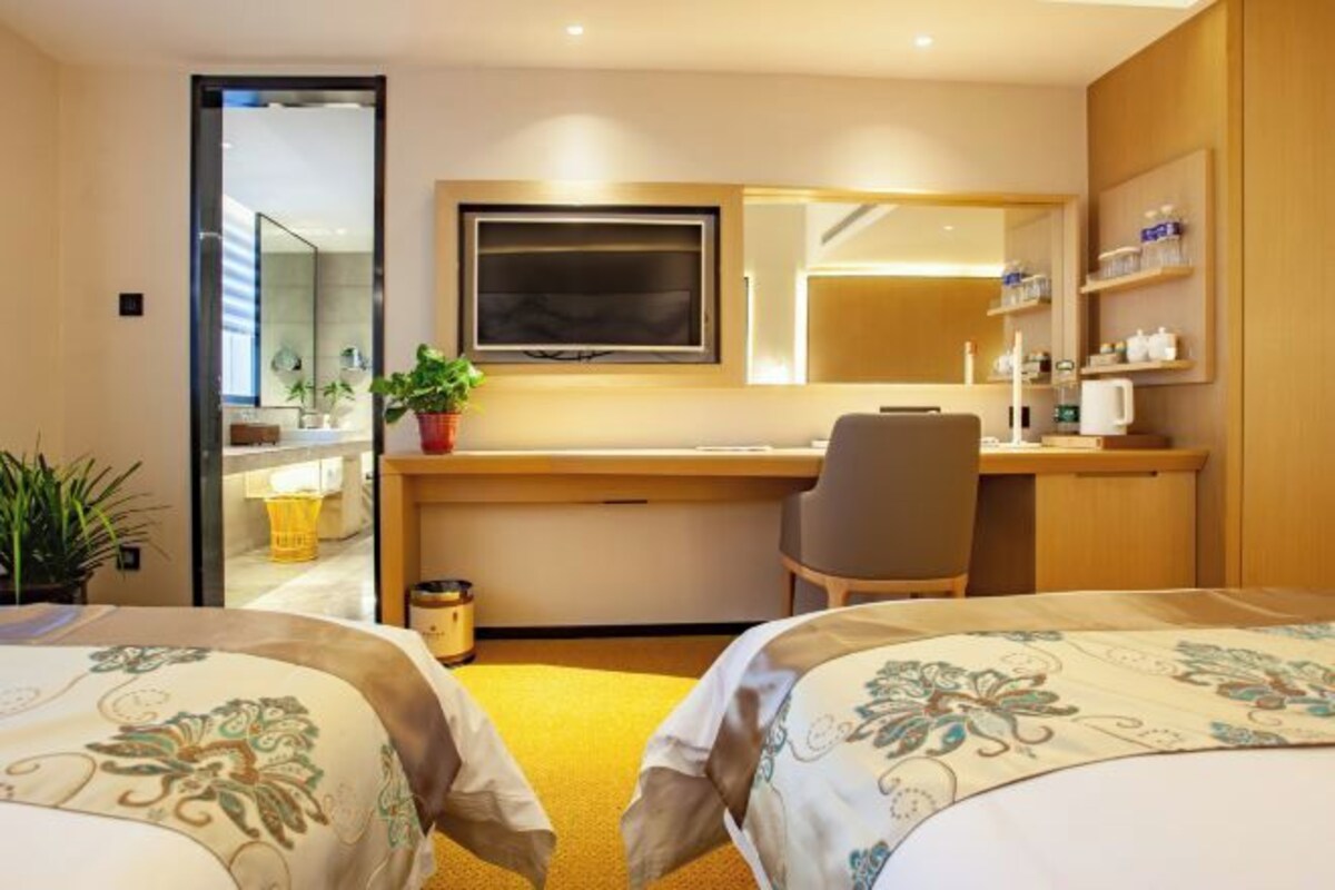 A hotel room with two beds, a desk, a chair, a TV, a mirror, and a potted plant.