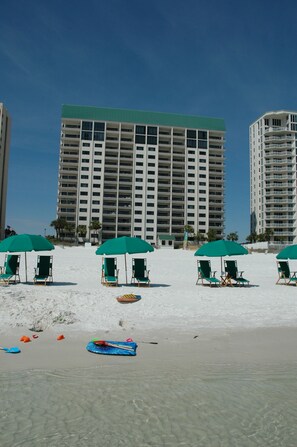A beach with multiple green umbrellas and lounge chairs, a large hotel building in the background, and clear blue skies.