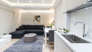 A modern living room with a dark grey sectional sofa, a round coffee table, and a dining area with a white table and chairs. A kitchen with a stainless steel sink and a white countertop is visible to the right. - 2 Palace Gardens (London)