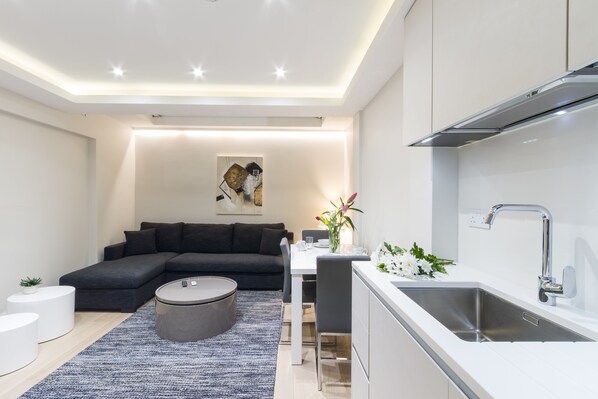 A modern living room with a dark grey sectional sofa, a round coffee table, and a dining area with a white table and chairs. A kitchen with a stainless steel sink and a white countertop is visible to the right. - 2 Palace Gardens (London)