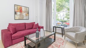 A modern living room with a red sofa, a glass coffee table, a grey armchair, and a large window with curtains. - 3b Imperial (London)