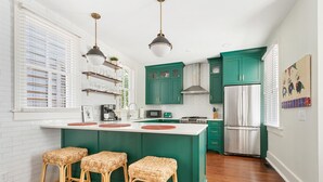 Una cocina con isla central, armarios verdes, electrodomésticos de acero inoxidable, taburetes de mimbre y una ventana con persianas.