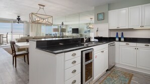 A modern kitchen with white cabinetry, a black countertop, and a large island with a sink. There is a dining area with a table and chairs, and a balcony with a view. - 2BR Beachfront Condo at Hidden Dunes! Beach Views, 3 Tropical Pools, Hot Tub. (Miramar Beach)