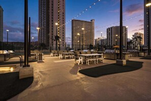 An outdoor seating area with tables and chairs, surrounded by tall buildings and string lights.