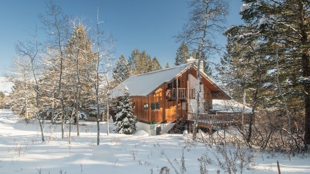 Lumihunnun peittämä puinen mökki, jonka ympärillä on puita ja lumipeitteinen maisema.