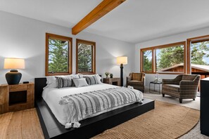 A modern bedroom with a large bed, two bedside lamps, a chair, and a view of trees through large windows.