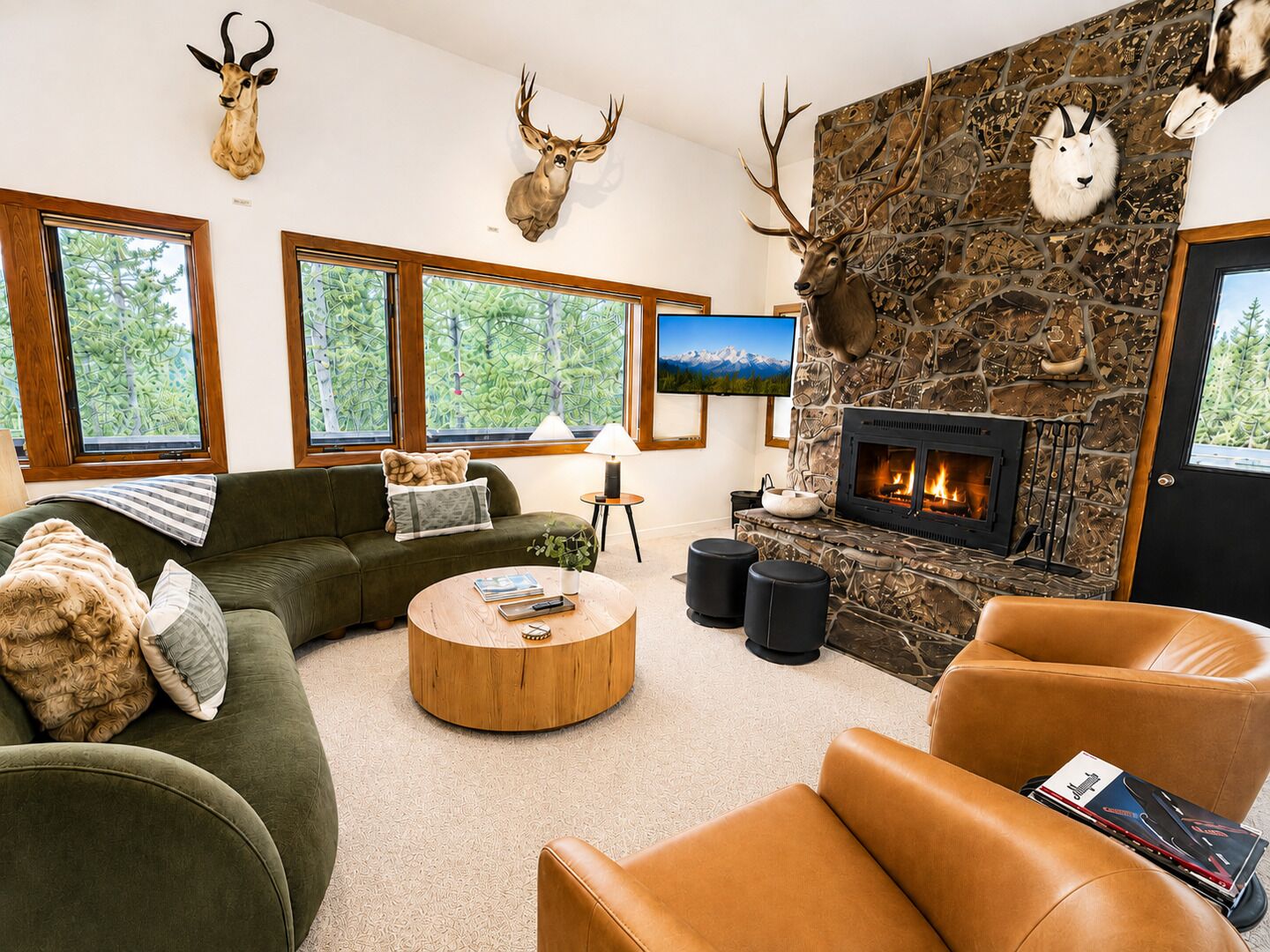 Un salón con chimenea de piedra, trofeos de animales en la pared, un sofá verde, sillones de cuero y un televisor.