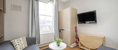 Ein modernes Wohnzimmer mit einer Couch, einem kleinen runden Tisch mit einer Pflanze, einem an der Wand befestigten Fernseher und einem Holzschrank.
