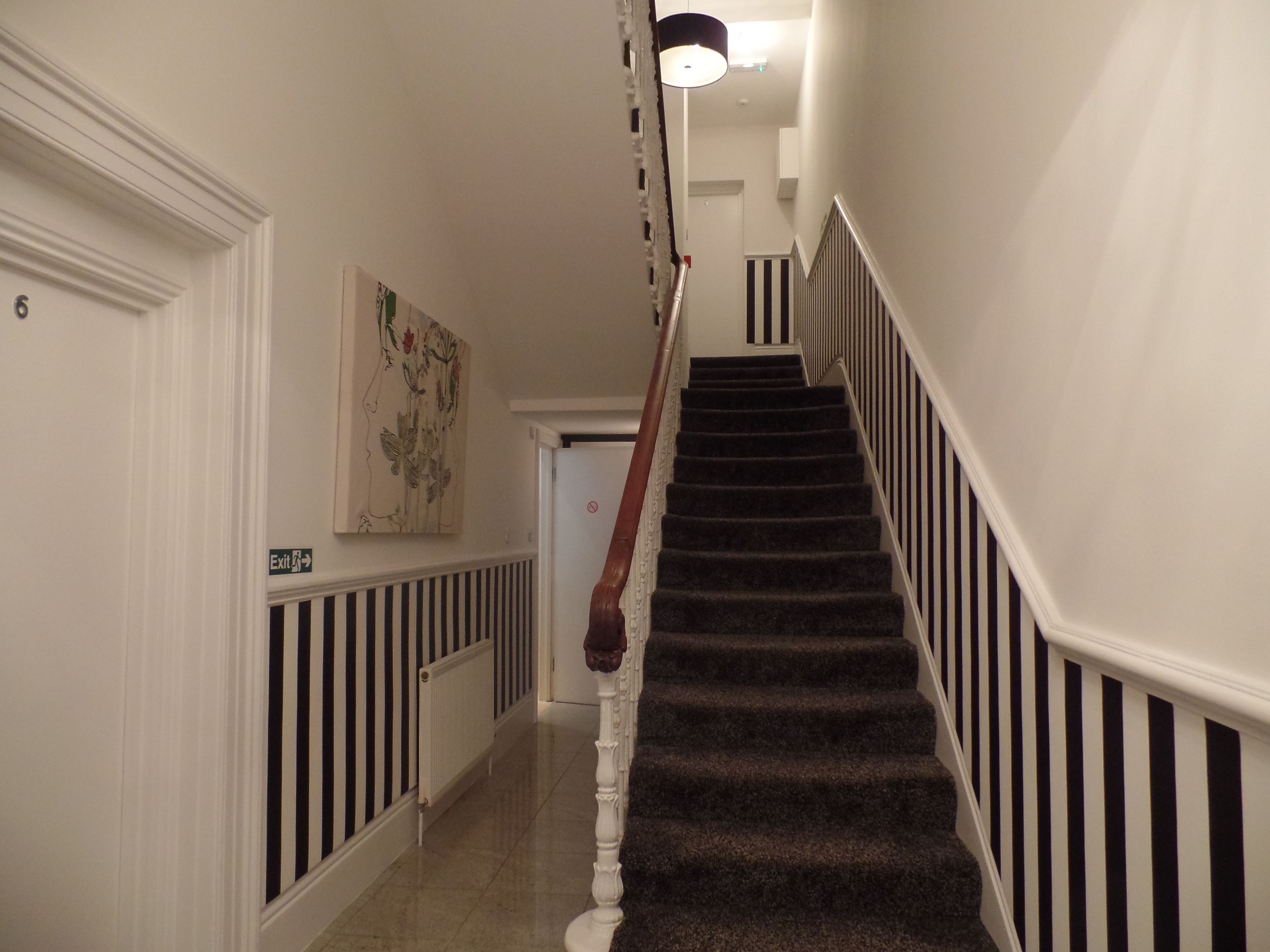 A staircase with black and white striped railings, a white door with a number '6' sign, and a wall-mounted exit sign.