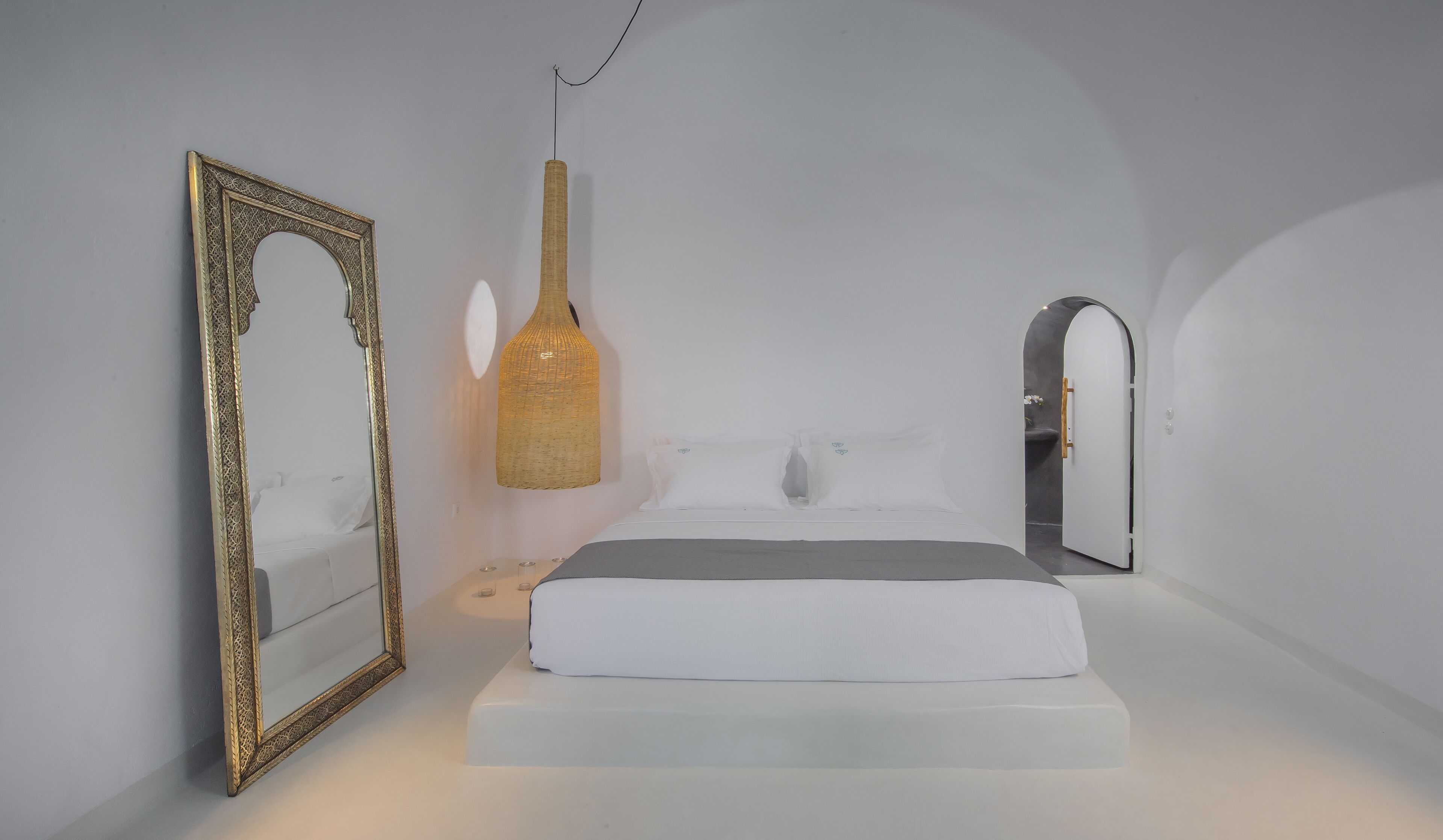 A minimalist bedroom with a large ornate mirror, a hanging light fixture, and a door leading to another room.