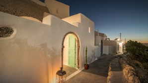 A narrow, stone-paved pathway leading to a white-walled building with a green door, a lantern, and a potted plant. - Sophia Finikia Villas (Santorini)