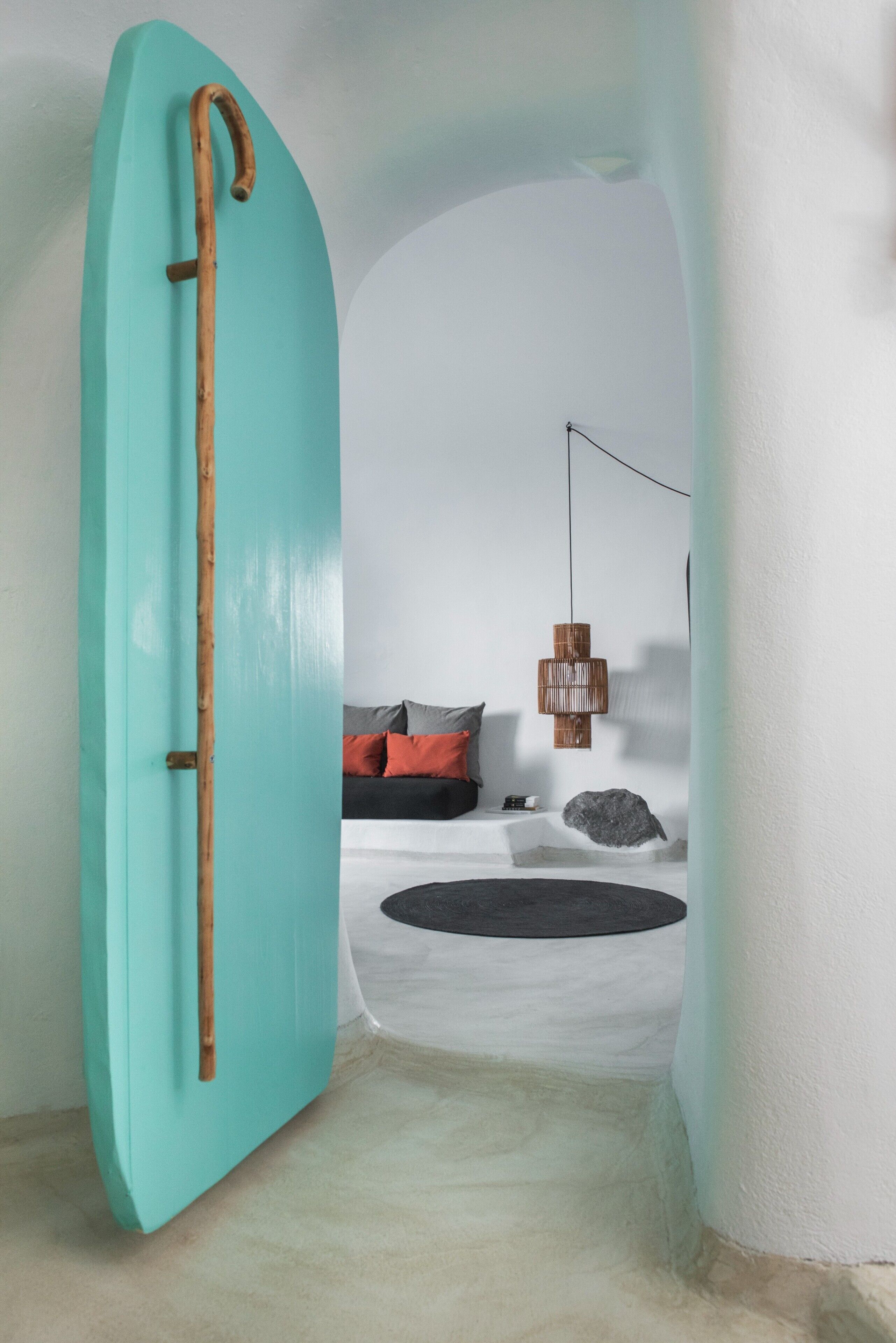 Una habitación con una tabla de surf color aguamarina apoyada en una pared, una cama con almohadas, una lámpara colgante y una piedra en el suelo.