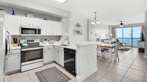 A modern kitchen with white cabinetry, a built-in microwave, and a dishwasher. There is a dining area with a table and chairs, and a view of the ocean through large windows.
