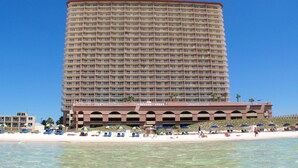 A large hotel with multiple balconies, a beach area with sun loungers and umbrellas, and clear blue water.