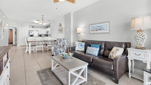 A spacious living area with a brown leather sofa, white coffee table, and patterned chairs. A ceiling fan and a dining area with white cabinets and a kitchen are visible in the background.
