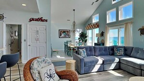 Un vaste espace de vie comprenant un grand canapé d’angle bleu, un fauteuil en osier agrémenté d’un coussin décoratif, ainsi qu’un coin repas avec une table et des chaises.