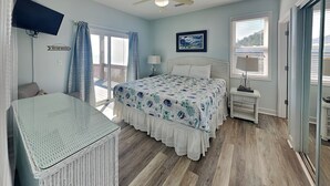 Une chambre à coucher comprenant un lit, un ventilateur de plafond, une télévision, une fenêtre avec des rideaux, une table de chevet et une chaise en osier.