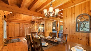 A wooden interior with a dining table set for six, surrounded by wooden chairs. A chandelier hangs from the ceiling, and a stone fireplace is visible in the background. - Large Cabin Near Downtown Blowing Rock (Blowing Rock)