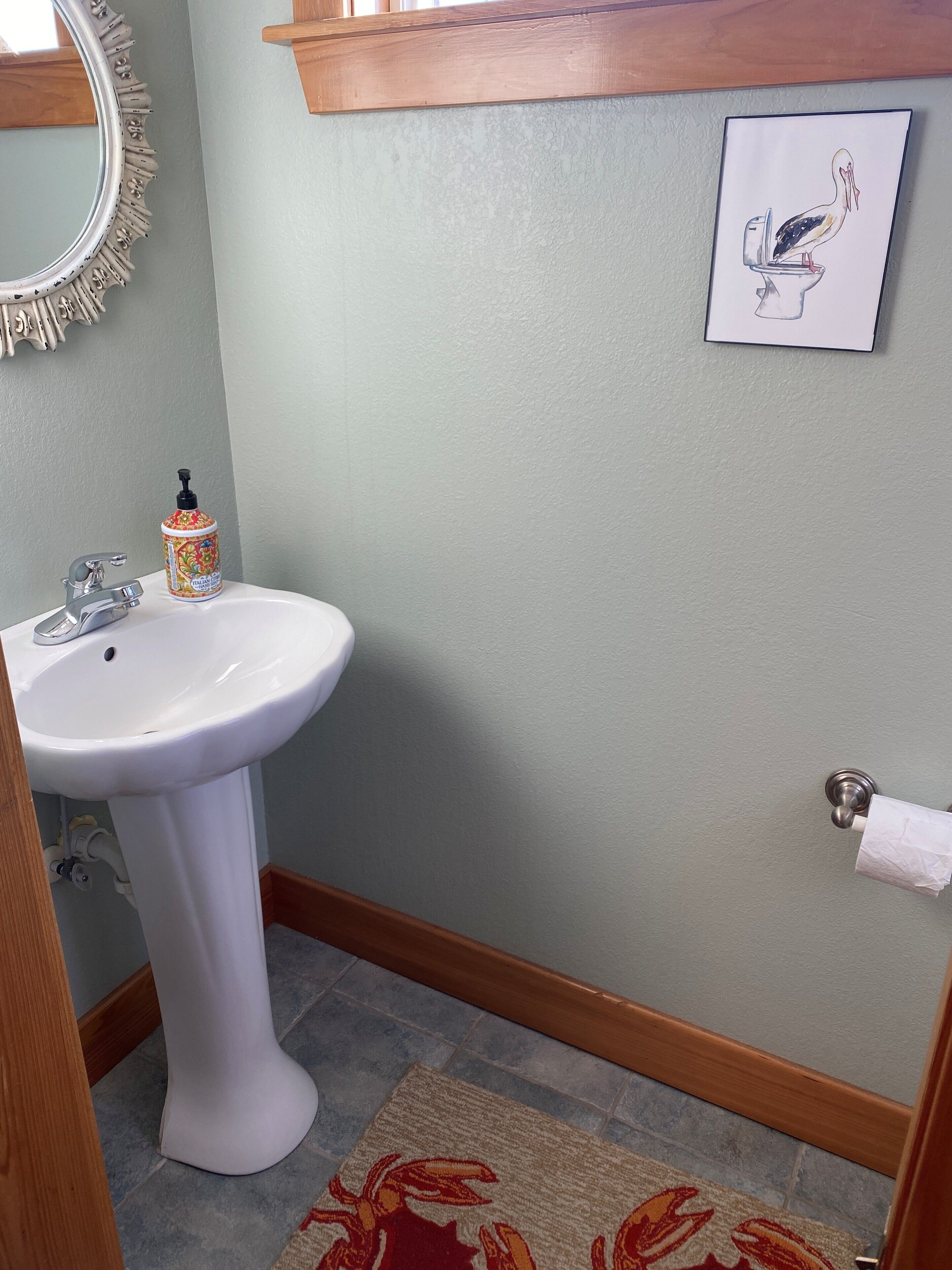 Et badeværelse med en hvid piedestalvask, et spejl med dekorativ ramme, en toiletpapirholder og en indrammet tegning af en fugl på toilettet.