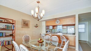 A dining area with a glass table surrounded by white chairs, a bookshelf, and a kitchen in the background.