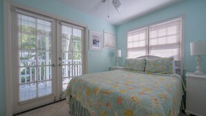 A bedroom with a bed, two bedside tables, a ceiling fan, and large windows with blinds. - Captain's Wheel at The Truman Annex in Key West (Key West)