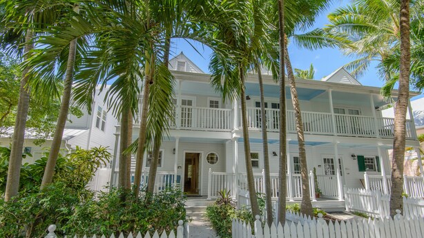 A two-story white house with a porch, surrounded by palm trees and a white picket fence. - Captain's Wheel at The Truman Annex in Key West (Key West)