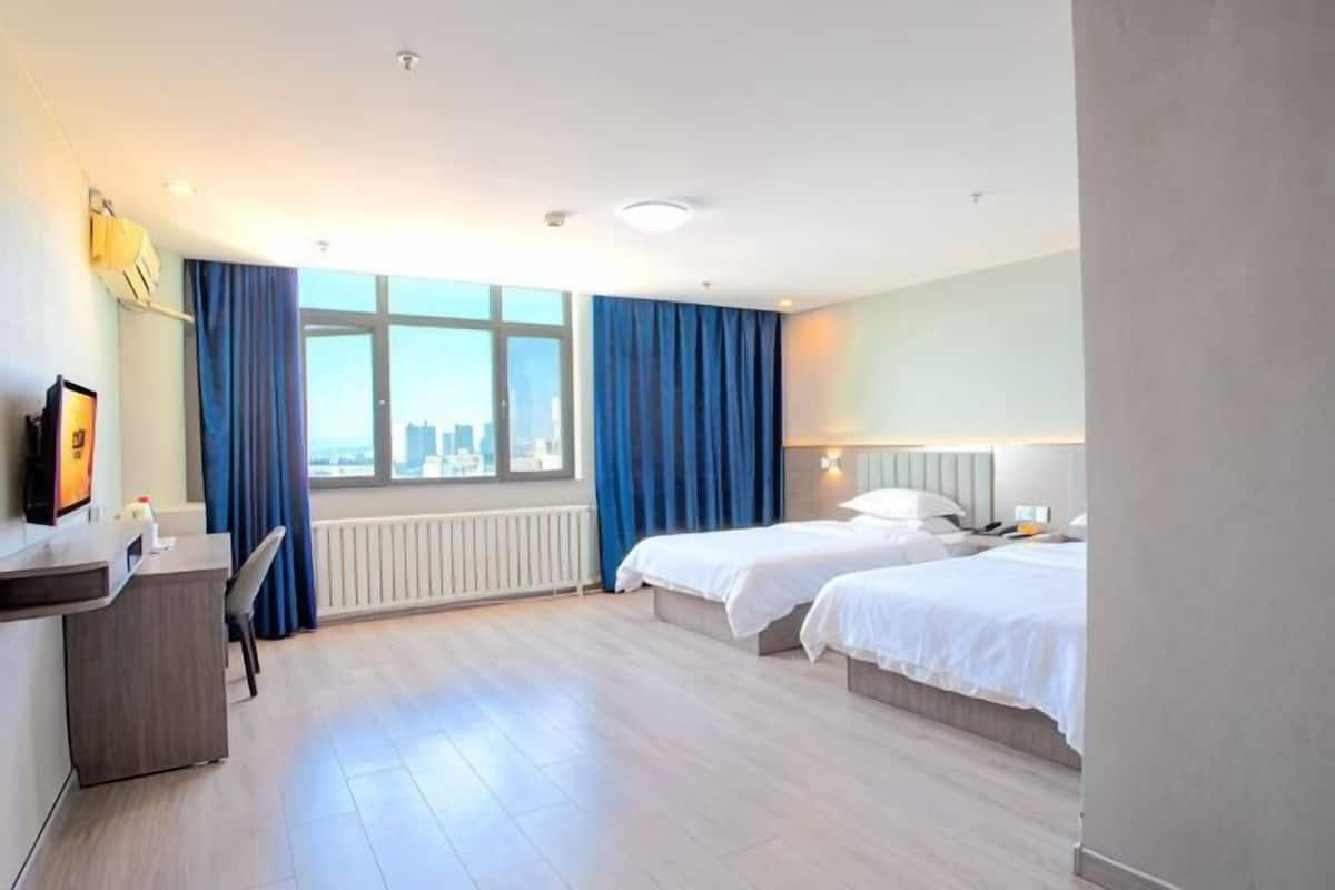 Habitación de hotel con dos camas, un escritorio, una silla, una ventana con cortinas y vistas a la ciudad.