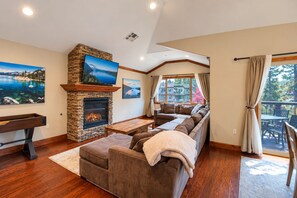 A living room with a stone fireplace, a large sectional sofa, a flat-screen TV mounted above the fireplace, and a view of a house through a large window.