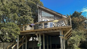 A two-story wooden house with a balcony, a staircase, and a covered outdoor area. - Bearfoot Bodega: Pet friendly on Back Road near the Coffee Co. (Ocracoke)