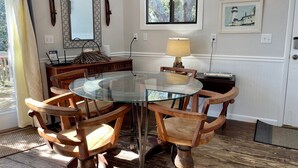 A dining area with a glass table and wooden chairs, a lamp, a framed picture, and a window with a view of trees. - Bearfoot Bodega: Pet friendly on Back Road near the Coffee Co. (Ocracoke)
